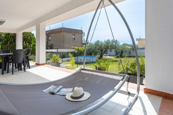 A hammock with a hat, a table with chairs, and a view of a pool and greenery.