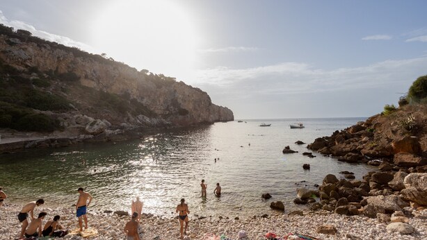 A rocky beach with clear water, a cliff, and boats in the distance.