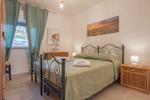 Ein Schlafzimmer mit einem Metallbettgestell, grüner Bettwäsche, einem Nachttisch mit Lampe, einem Weidenholz-Nachttisch, einem Sonnenuntergangsbild und einem Fenster mit Vorhängen.