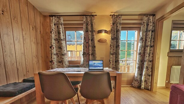 A dining area with a wooden table, two chairs, a laptop, and a view of a balcony. - Holiday Apartment Chalet Monteneve Toilà with Mountain View, Balcony & Wi-Fi (livigno)