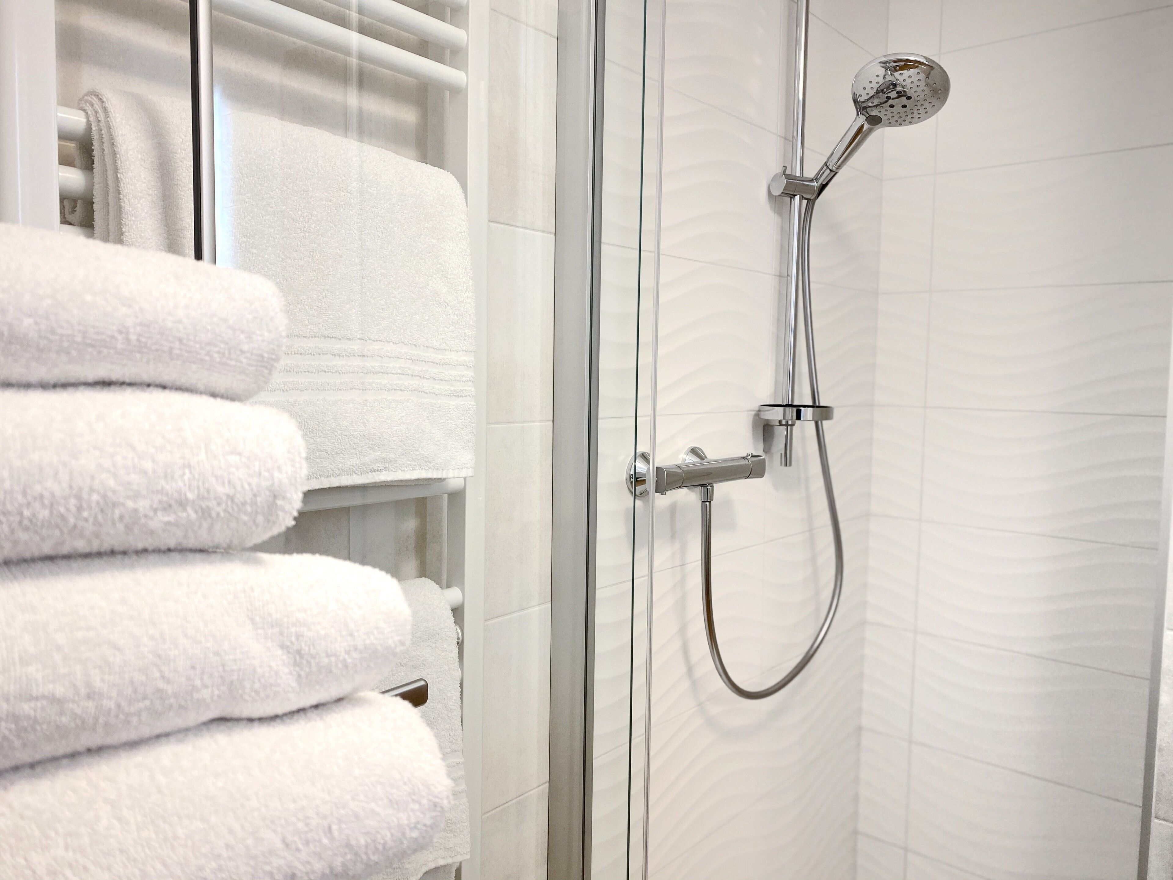 A shower with a handheld showerhead and a wall-mounted faucet, a glass shower enclosure, and a stack of white towels on a rack.