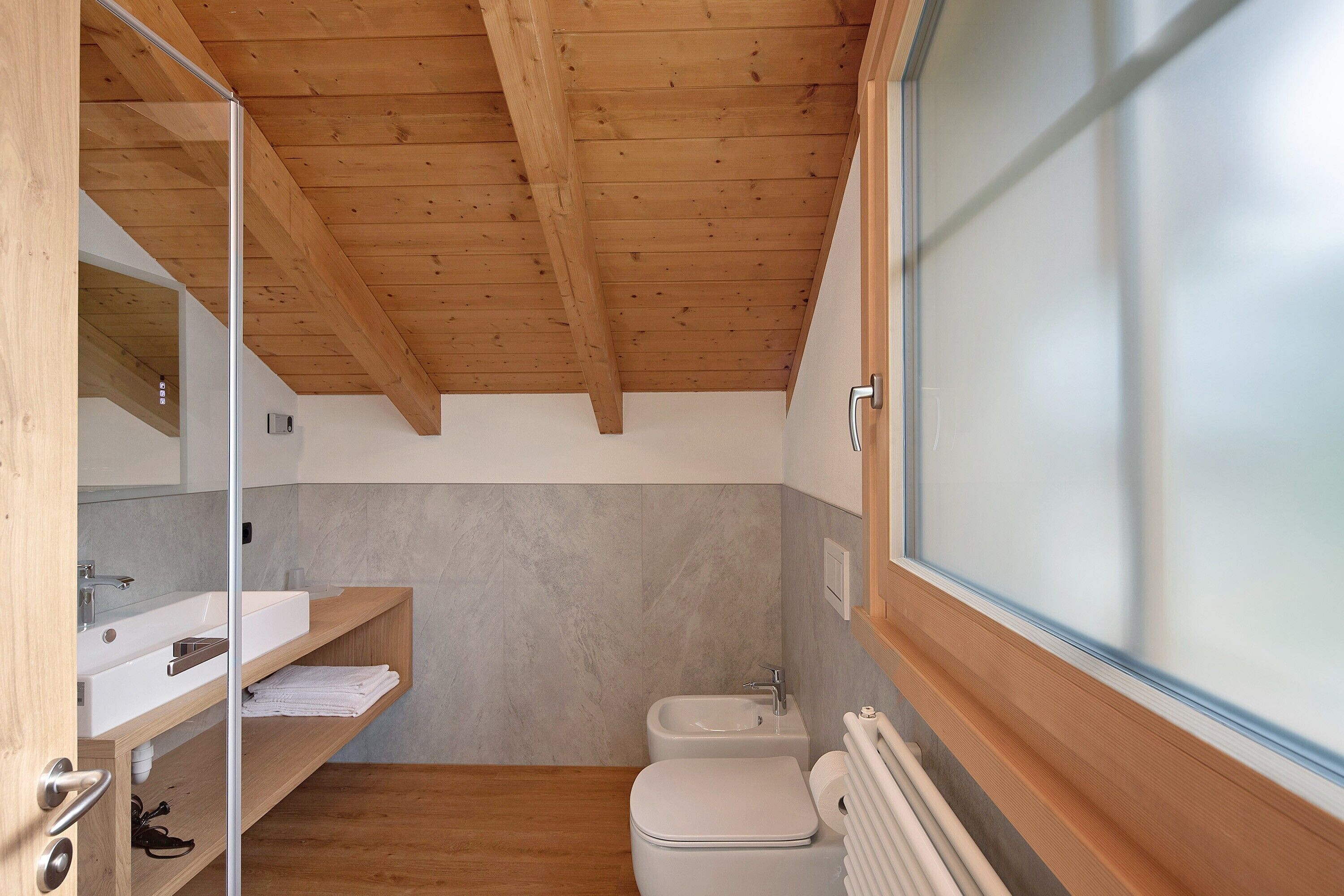 Ein modernes Badezimmer mit Holzboden, einem weißen Waschbecken, einer Toilette und einem Fenster mit satinierten Scheiben.