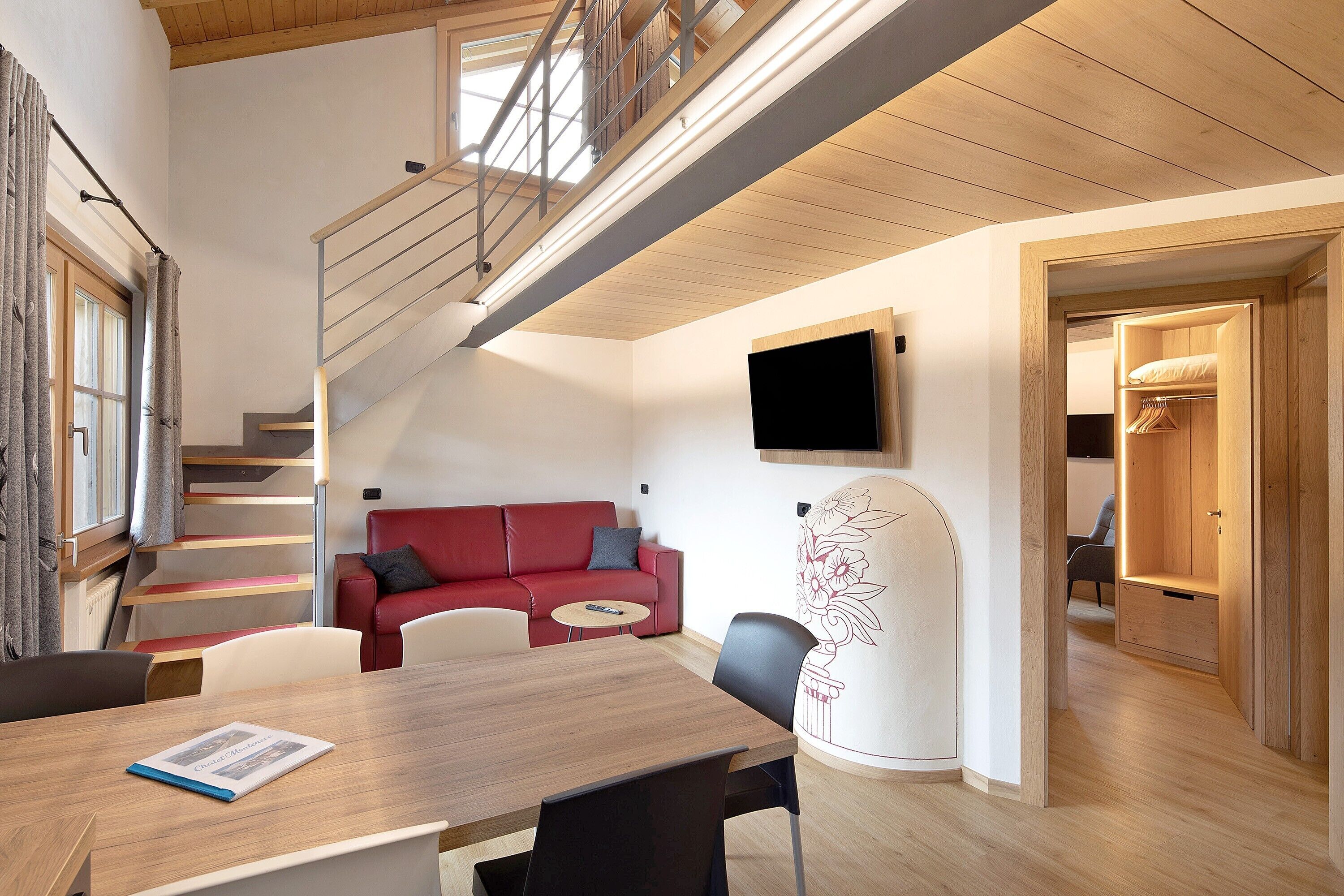 Un interior moderno con comedor, sofá rojo, escalera y televisor montado en la pared.