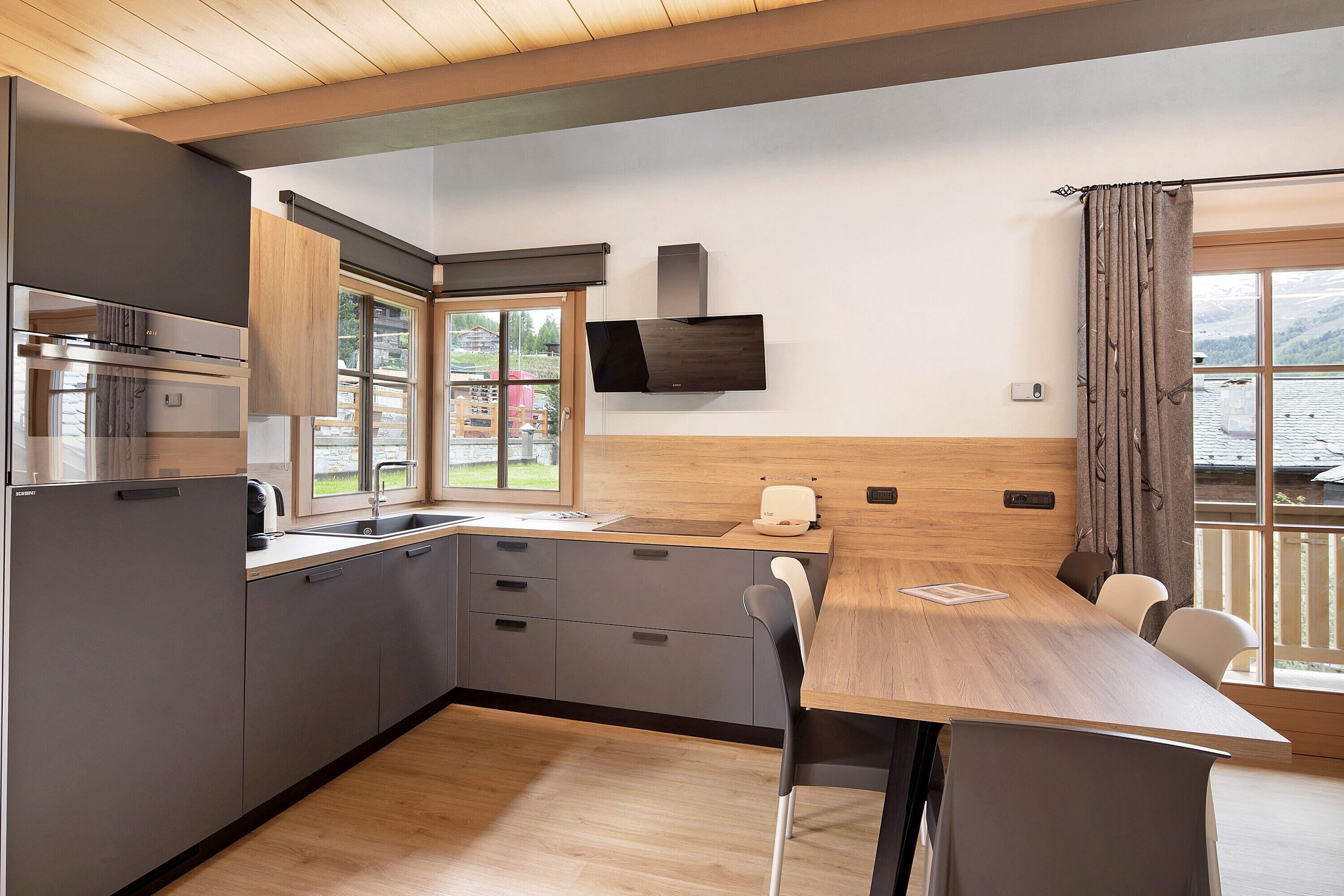 A modern kitchen with a wooden table, built-in appliances, and large windows.