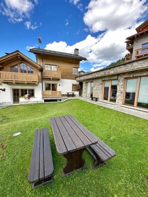 Une maison à deux étages avec une façade en pierre, des balcons en bois et une cour verdoyante équipée de tables de pique-nique.
