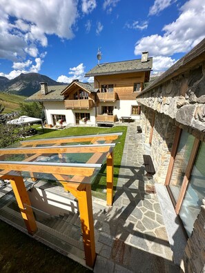 Un chalet con tavolo in vetro, pareti in pietra e vista sulle montagne.