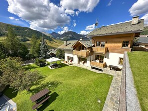 Une maison en bois à deux étages, dotée d’un balcon, d’une cheminée en pierre et offrant une vue sur un paysage montagneux.