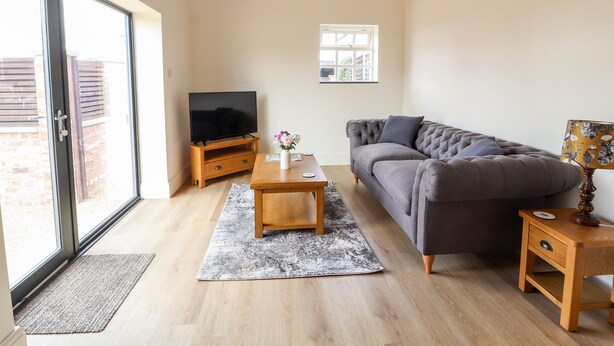 Un salon moderne avec un plafond en bois, un canapé gris, une table basse en bois, un téléviseur posé sur un meuble en bois et une petite table de chevet en bois avec une lampe.