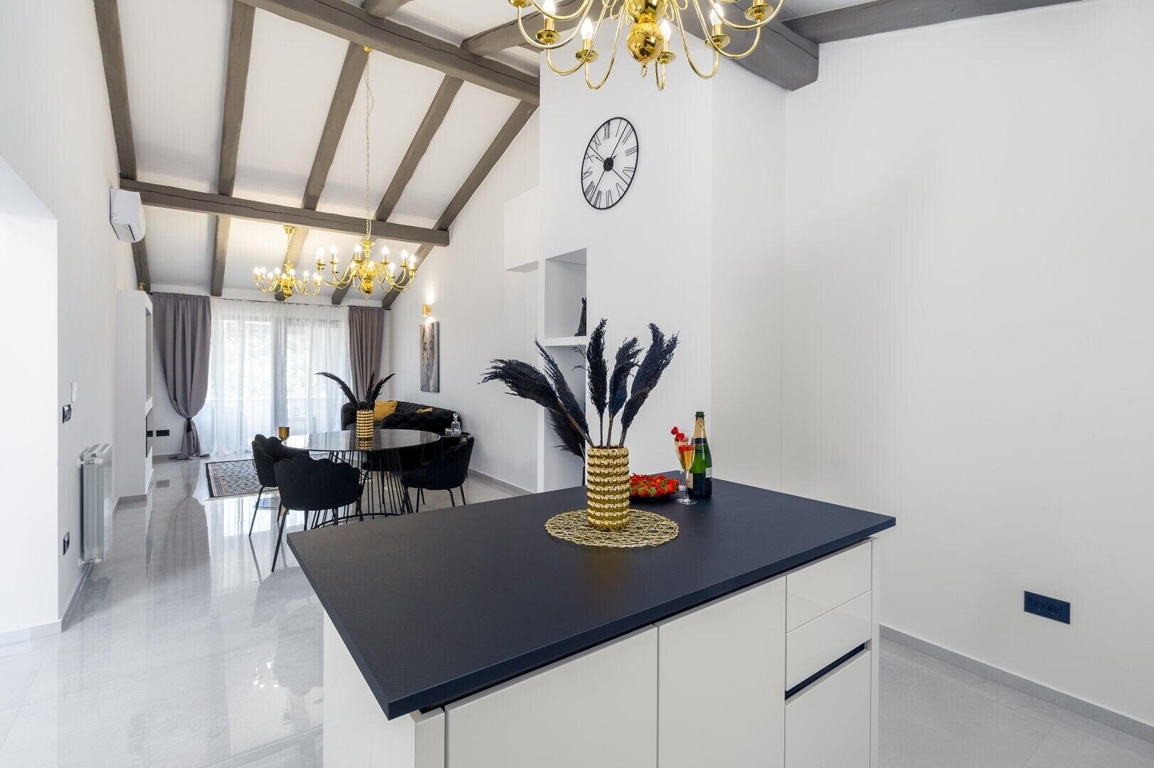 A modern kitchen with a black countertop, white cabinets, and a chandelier. There is a dining area with a table and chairs, and a clock on the wall.