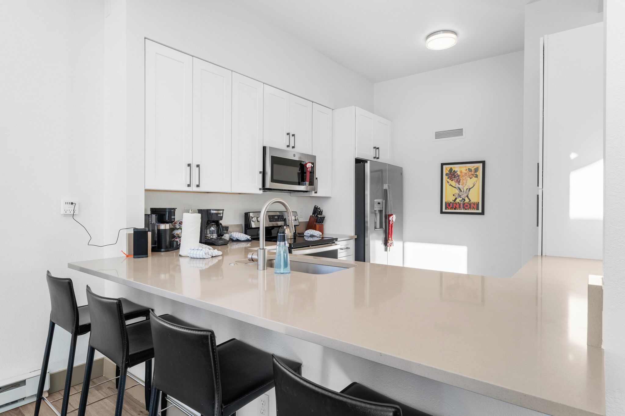A modern kitchen with white cabinets, a stainless steel refrigerator, a microwave, and a coffee machine. There are black bar stools at the island.