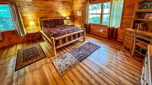 A wooden cabin bedroom with a bed, two nightstands, a ceiling fan, and a bookshelf. - Breezy River Cabin | Mountain Views, Hot Tub, Game Room & River Access (Piney Creek)