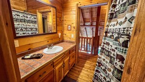 A wooden cabin bathroom with a sink, mirror, and patterned shower curtain. - Breezy River Cabin | Mountain Views, Hot Tub, Game Room & River Access (Piney Creek)