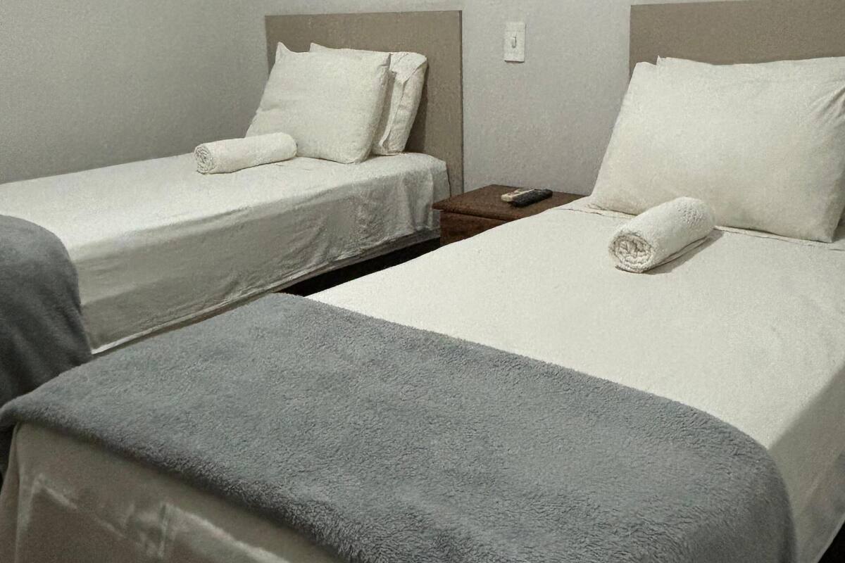 Two single beds with white bedding and grey blankets, a wooden nightstand, and a plain wall.