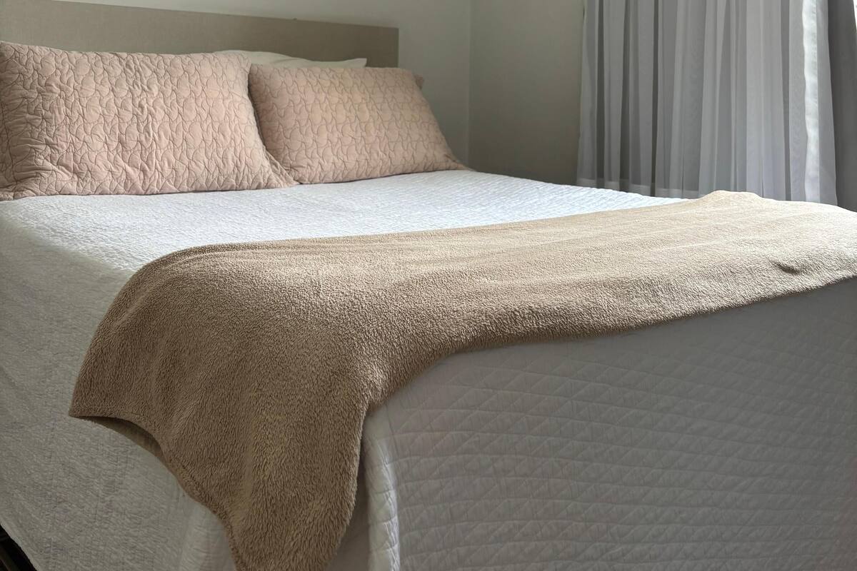 A neatly made bed with a beige blanket and pillows, a white quilted bedspread, and a sheer curtain partially drawn to the side.