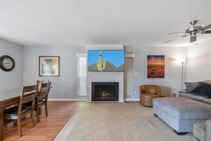 Una sala de estar con chimenea, un televisor montado encima, un comedor con sillas, un sofá y un ventilador de techo.