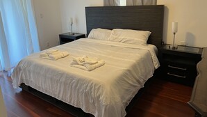 Una cama bien tendida con ropa blanca y toallas, un cabecero oscuro y una mesita de noche con una lámpara.