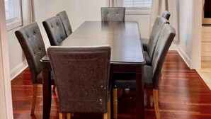 Comedor con una mesa rectangular, seis sillas tapizadas, una araña, un reloj y un ventanal con persianas.