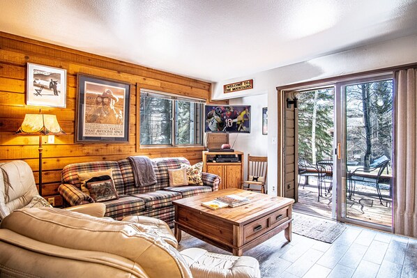 A cozy living room with a plaid sofa, a wooden coffee table, and a television. There are framed pictures on the wall and a sign that reads "Welcome to the Mountains." - Elkhorn Bluff Condo with Community & Resort Amenities (Sun Valley)