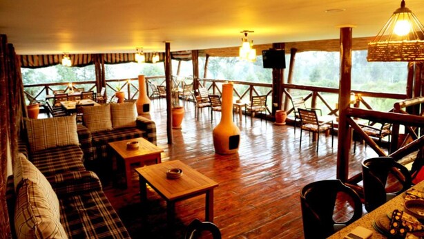 A cozy indoor seating area with wooden tables and sofas, surrounded by large windows offering a view of the outdoors. - Da Vinci Gorilla Lodge (Muhoza)