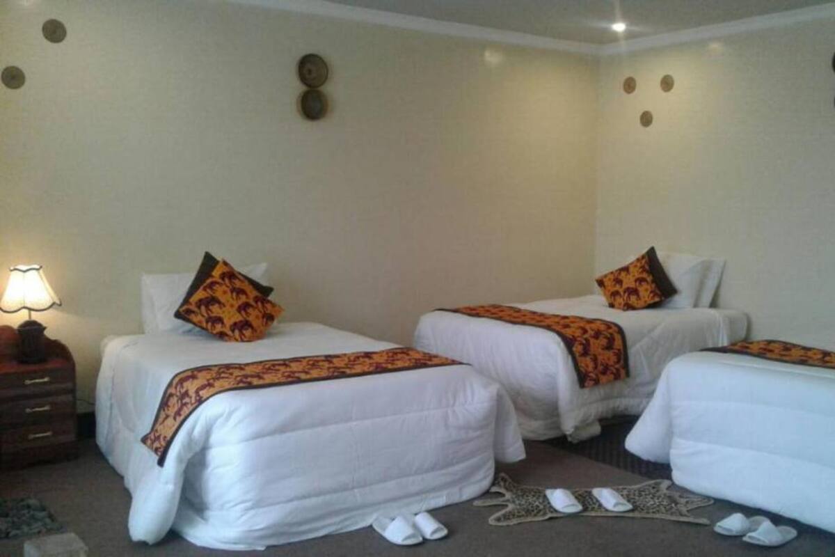 Habitación de hotel con tres camas individuales, cada una con edredón blanco y manta decorativa estampada. Se ve un buró con una lámpara y una mesita con un objeto decorativo.