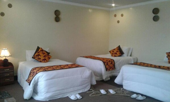 A hotel room with three single beds, each with a white duvet and a patterned throw blanket. There is a nightstand with a lamp and a small table with a decorative item.