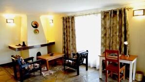 A cozy room with a stone floor, a wooden table, two chairs, a small shelf, and a lamp. - Da Vinci Gorilla Lodge (Muhoza)