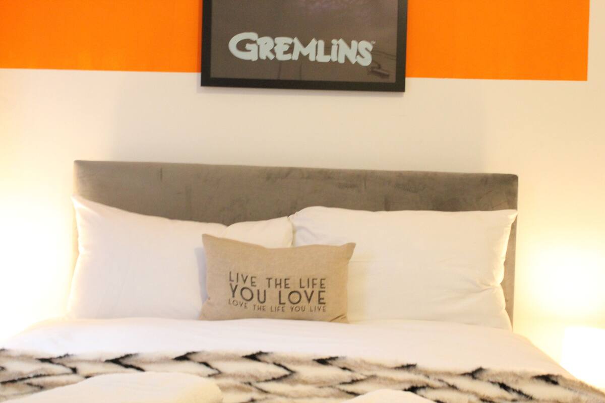 Una cama bien tendida con un cojín que dice 'Vive la vida que amas' y un cartel enmarcado encima que dice 'GREMLINS'.