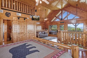 A cabin interior with a large bed, a sofa, a TV mounted on the wall, and a balcony with a view of the mountains. - Luxurious Cabin|Majestic Views|Arcade|Hot Tub (Sevierville)