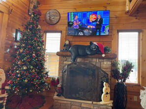 A cozy cabin interior with a decorated Christmas tree, a stone fireplace, and a television displaying a show. - Luxurious Cabin|Majestic Views|Arcade|Hot Tub (Sevierville)