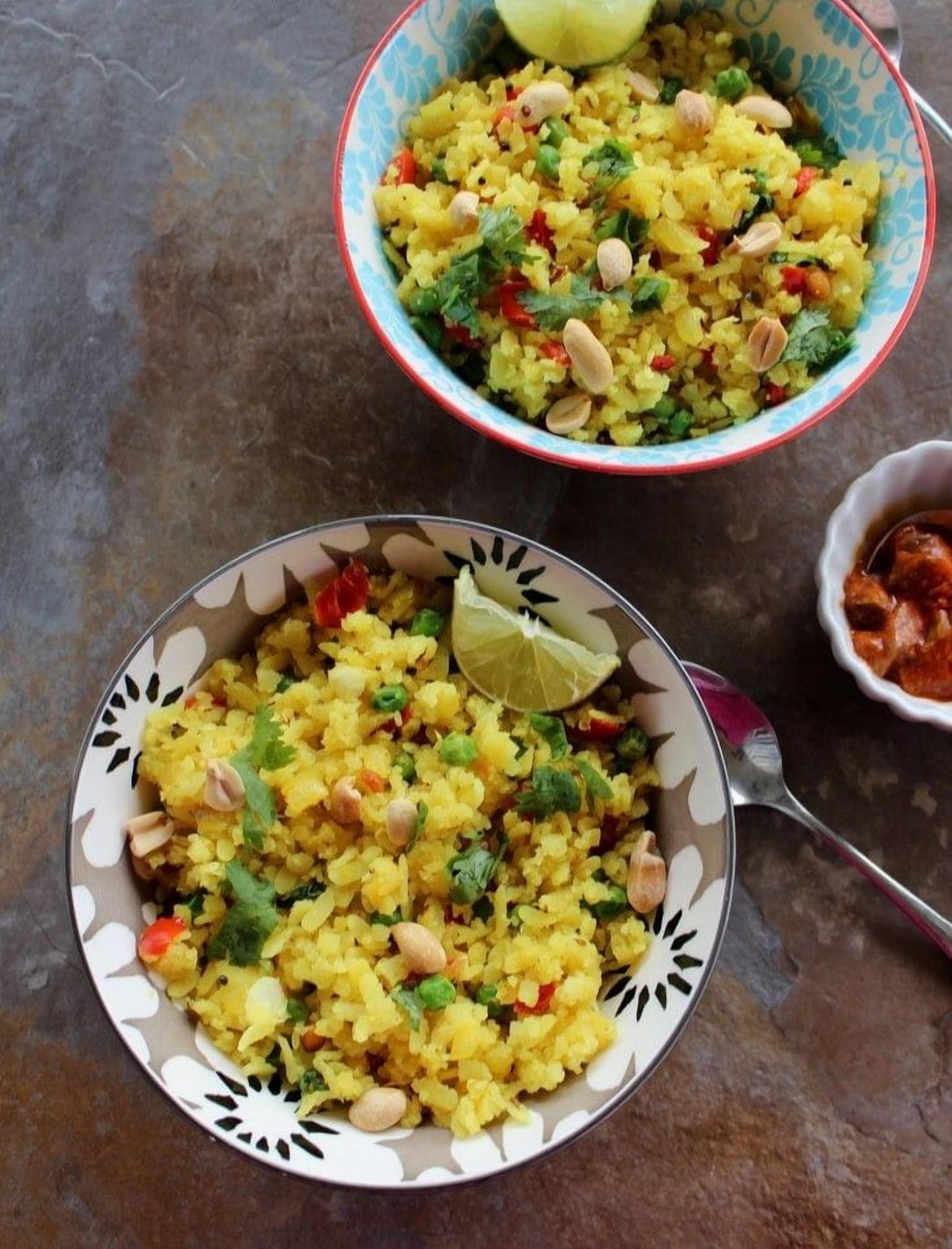 Dos tazones de Poha Upma, uno con una cuña de lima, uno sobre un plato estampado y otro sobre un plato liso.