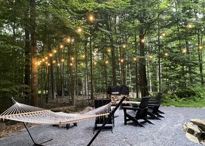 An outdoor seating area with a hammock, tables, and chairs, surrounded by trees and string lights.