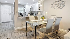 Cozinha moderna com armários brancos, micro-ondas e cafeteira. Possui um espaço de refeições com mesa de vidro e cadeiras.