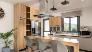 A modern kitchen with wooden cabinets, a central island with bar stools, stainless steel appliances, and a large window offering a view. - Mirena Luxury Living - Casa di Maria (Αφάντου)