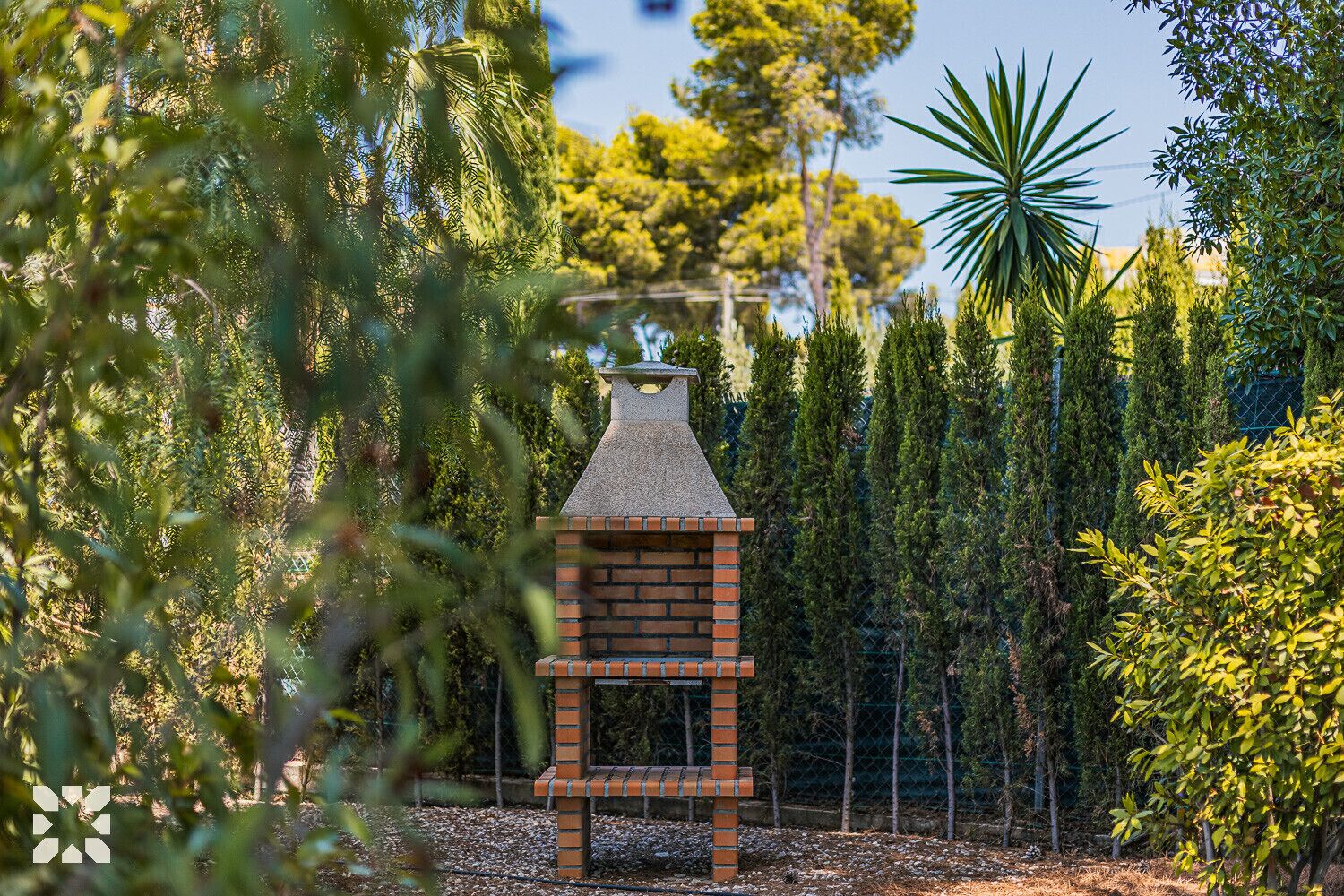 A brick-built outdoor barbecue grill surrounded by dense greenery and tall trees.