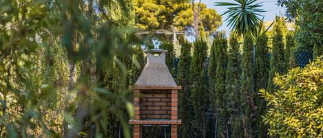 A brick-built outdoor barbecue grill surrounded by dense greenery and tall trees.