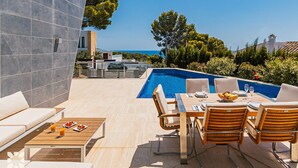A poolside area with a table set for dining, a sofa, and a wooden coffee table with fruit. - Villa ALEX by Abahana Villas (Benissa)