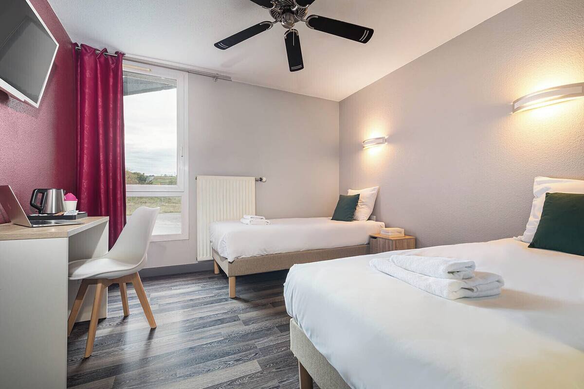 A hotel room with two beds, a desk with a laptop, a chair, a ceiling fan, and a window with curtains.
