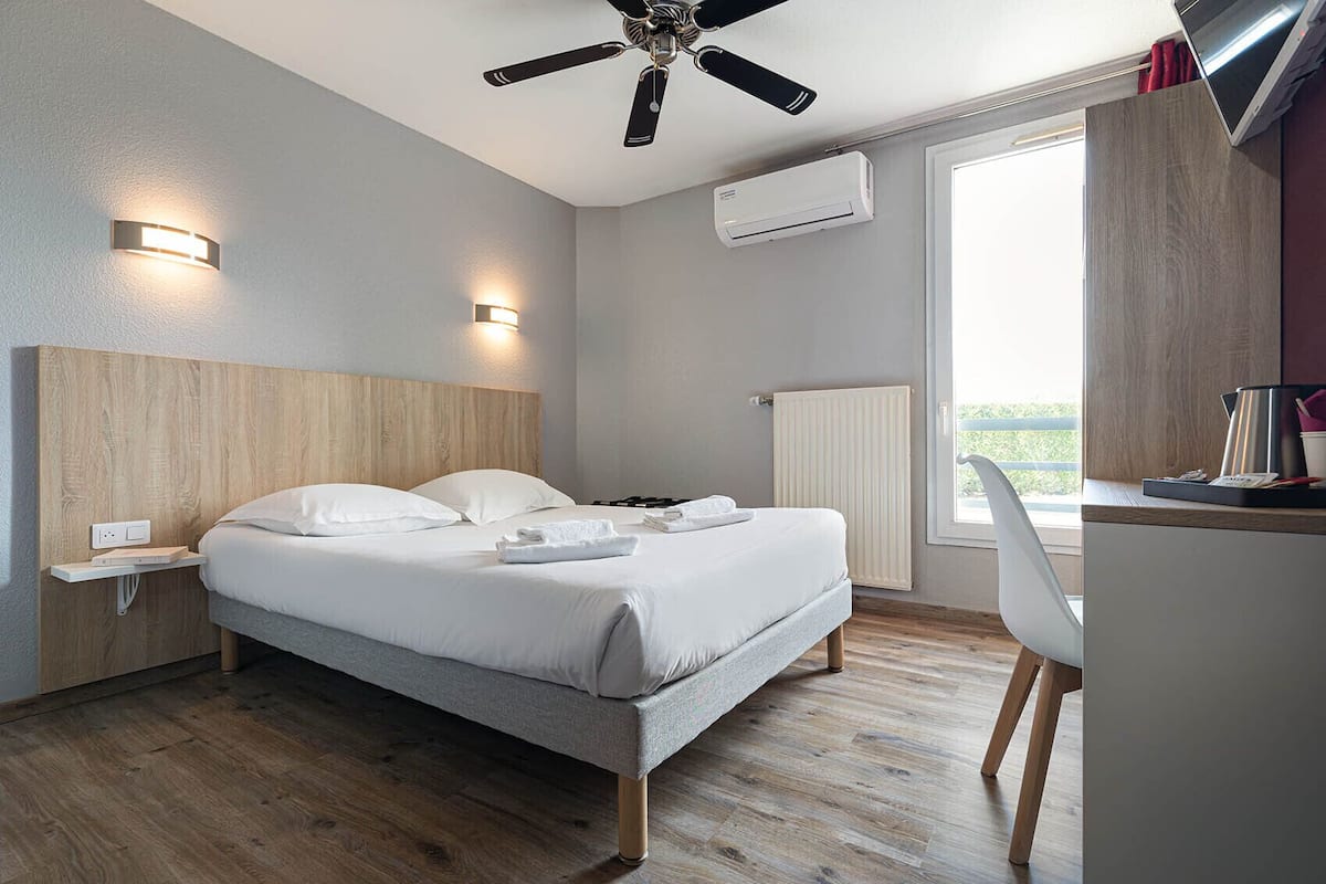 A modern hotel room with a wooden headboard, a bed with white linens, a desk with a chair, a ceiling fan, and an air conditioning unit.