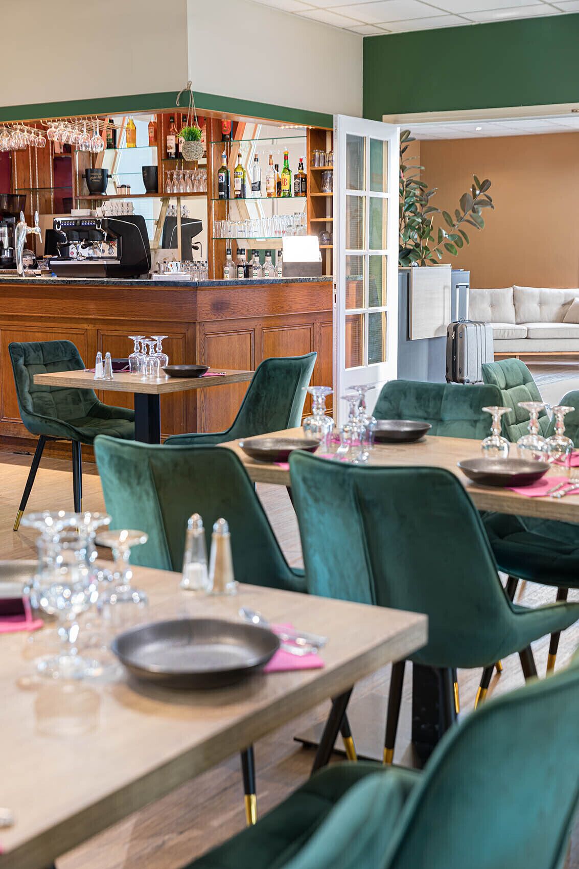 Un ristorante con tavoli di legno e sedie verdi, una zona bar con bottiglie e bicchieri, e un divano sullo sfondo.