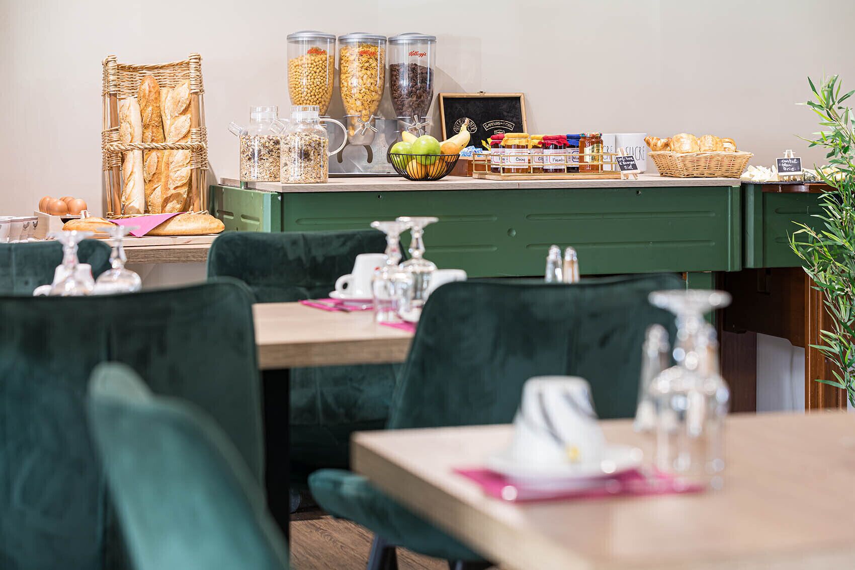 A restaurant interior with green upholstered chairs, a wooden table, and a counter with various food items and condiments.