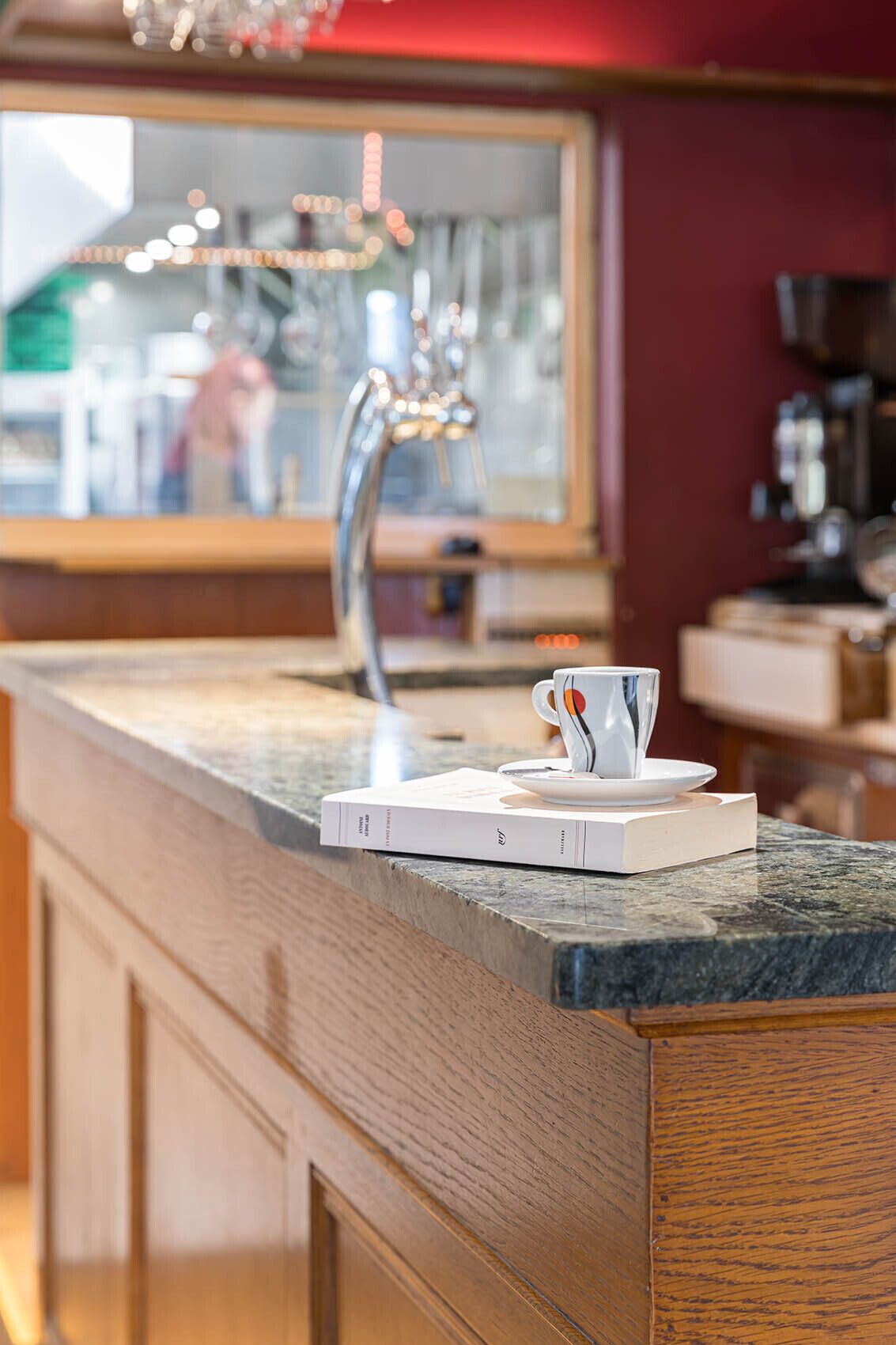 A kitchen counter with a coffee cup and a book, a stainless steel faucet, and a window in the background.