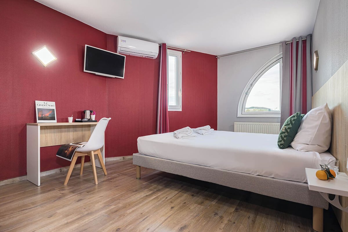 A modern hotel room with a bed, a desk, a chair, a TV, and a window with curtains.