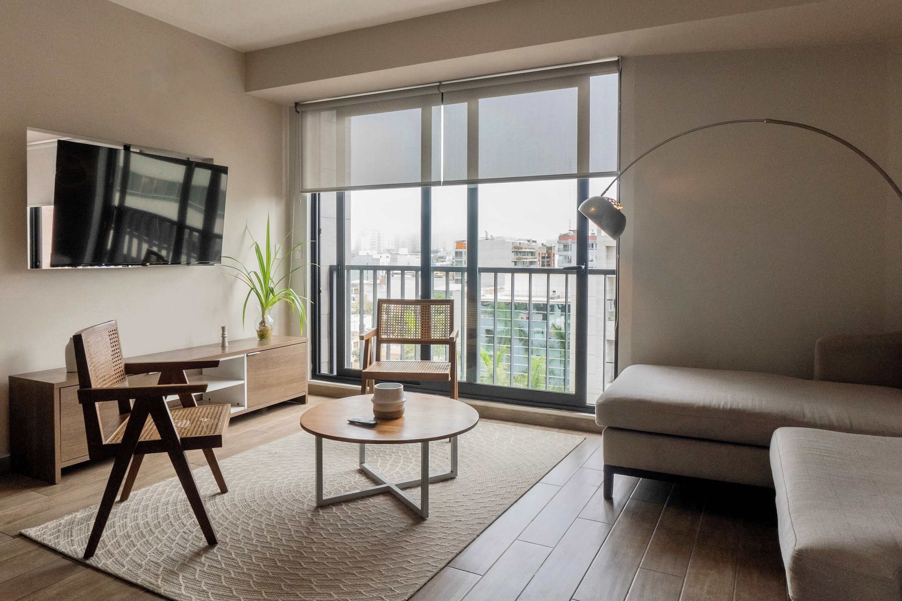 A modern living room with a sofa, a coffee table, a TV mounted on the wall, and a large window offering a city view.