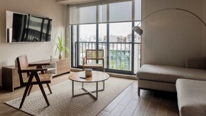 A modern living room with a sofa, a coffee table, a TV mounted on the wall, and a large window offering a city view.