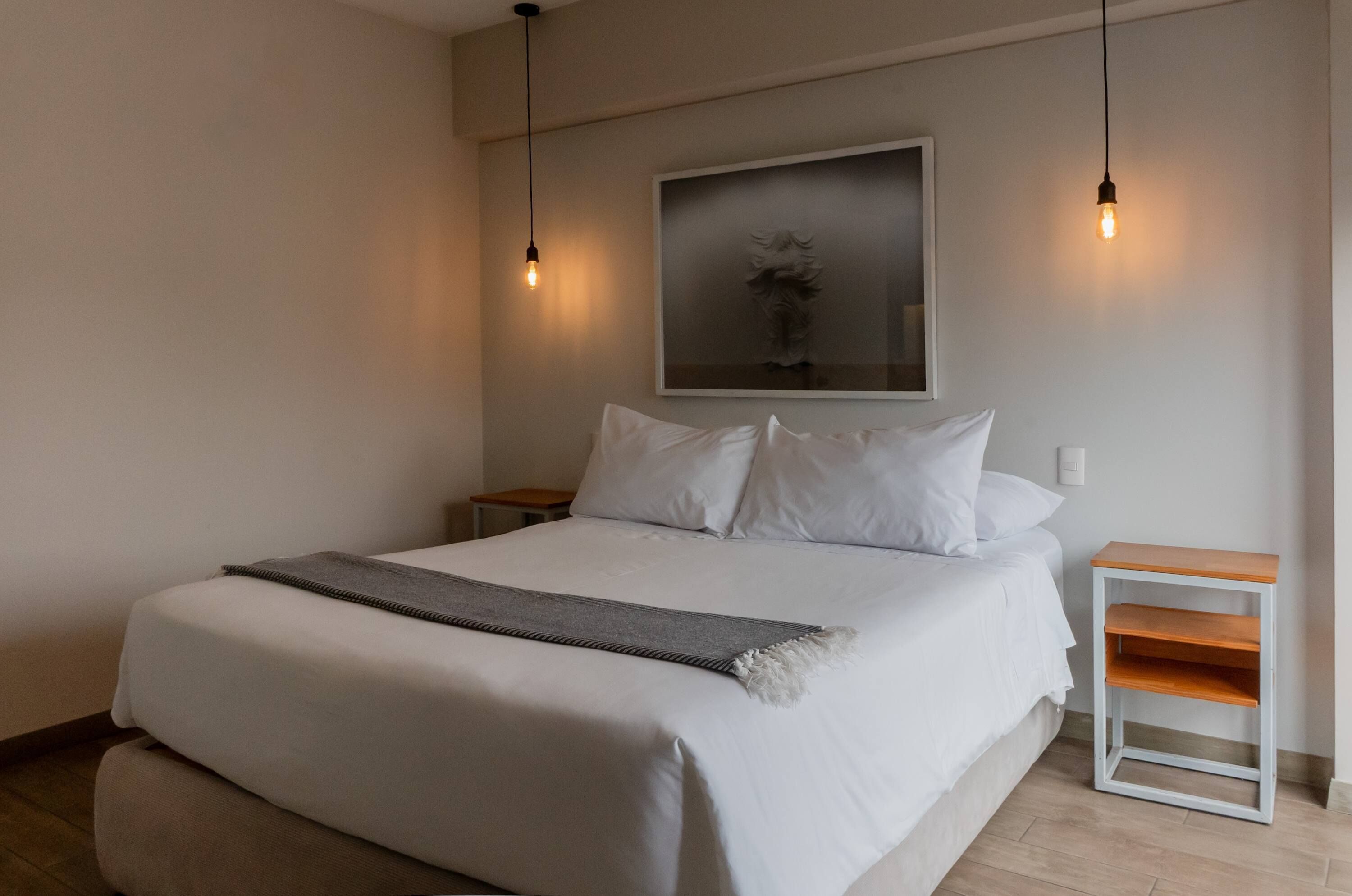 A neatly made bed with white linens and a gray throw blanket, a small wooden nightstand, and a wall-mounted light fixture.