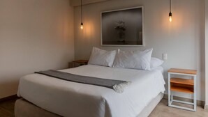 A neatly made bed with white linens and a gray throw blanket, a small wooden nightstand, and a wall-mounted light fixture.