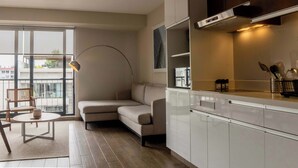 A modern living room with a beige sectional sofa, a round coffee table, and a floor lamp. There is a large window with a view of buildings outside.