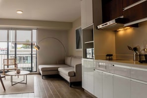 Un salon moderne avec un canapé d’angle beige, une table basse ronde et un lampadaire. Une grande fenêtre offre une vue sur les bâtiments environnants.