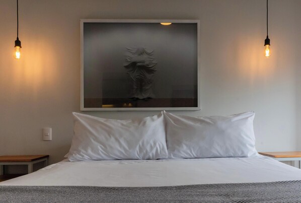 Habitación de hotel con una cama, dos mesitas de noche, una lámpara fijada a la pared y una obra de arte enmarcada sobre la cama.