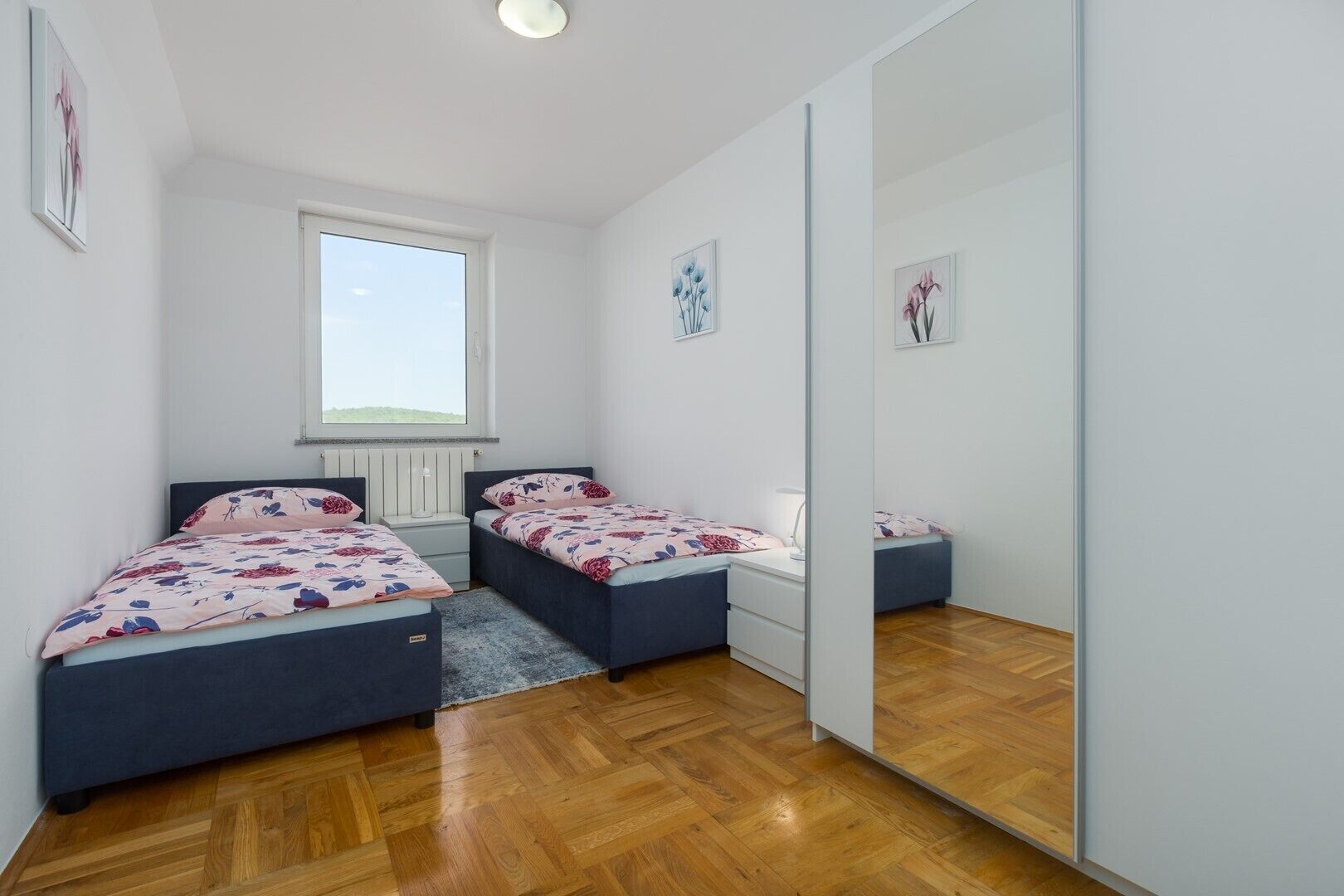 A bedroom with two beds, a large window, a mirror, and a radiator.